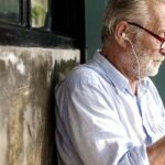 Man sitting on a bench on a sidewalk listening to something in headphones from his phone